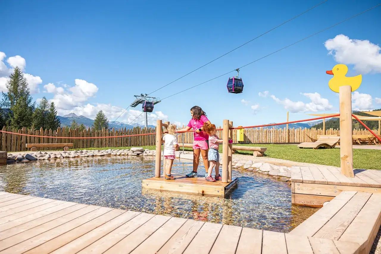 Bambini giocano in un parco acquatico a Cavalese, con funivie sullo sfondo.