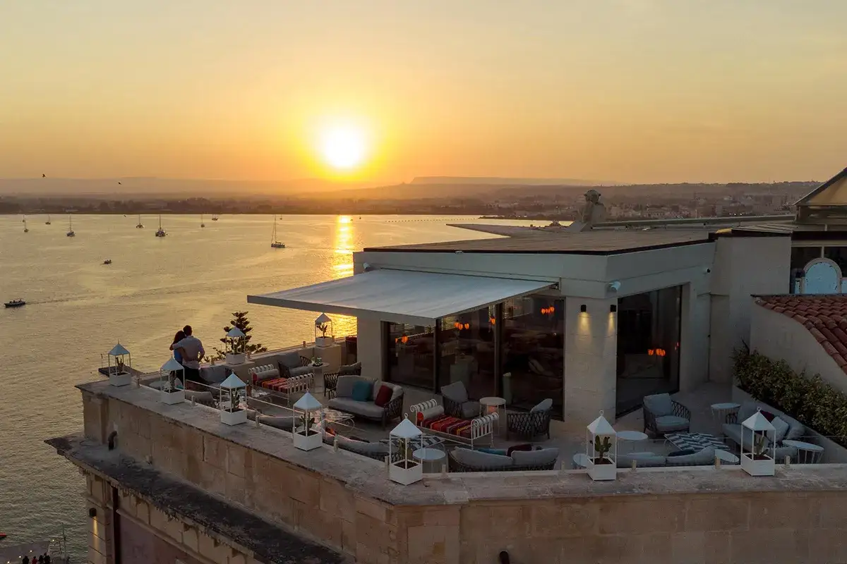 Terrazza panoramica con arredi eleganti e vista sul mare al tramonto. Coppia abbracciata ammira il sole che scende, un'atmosfera da sogno in questo hotel.