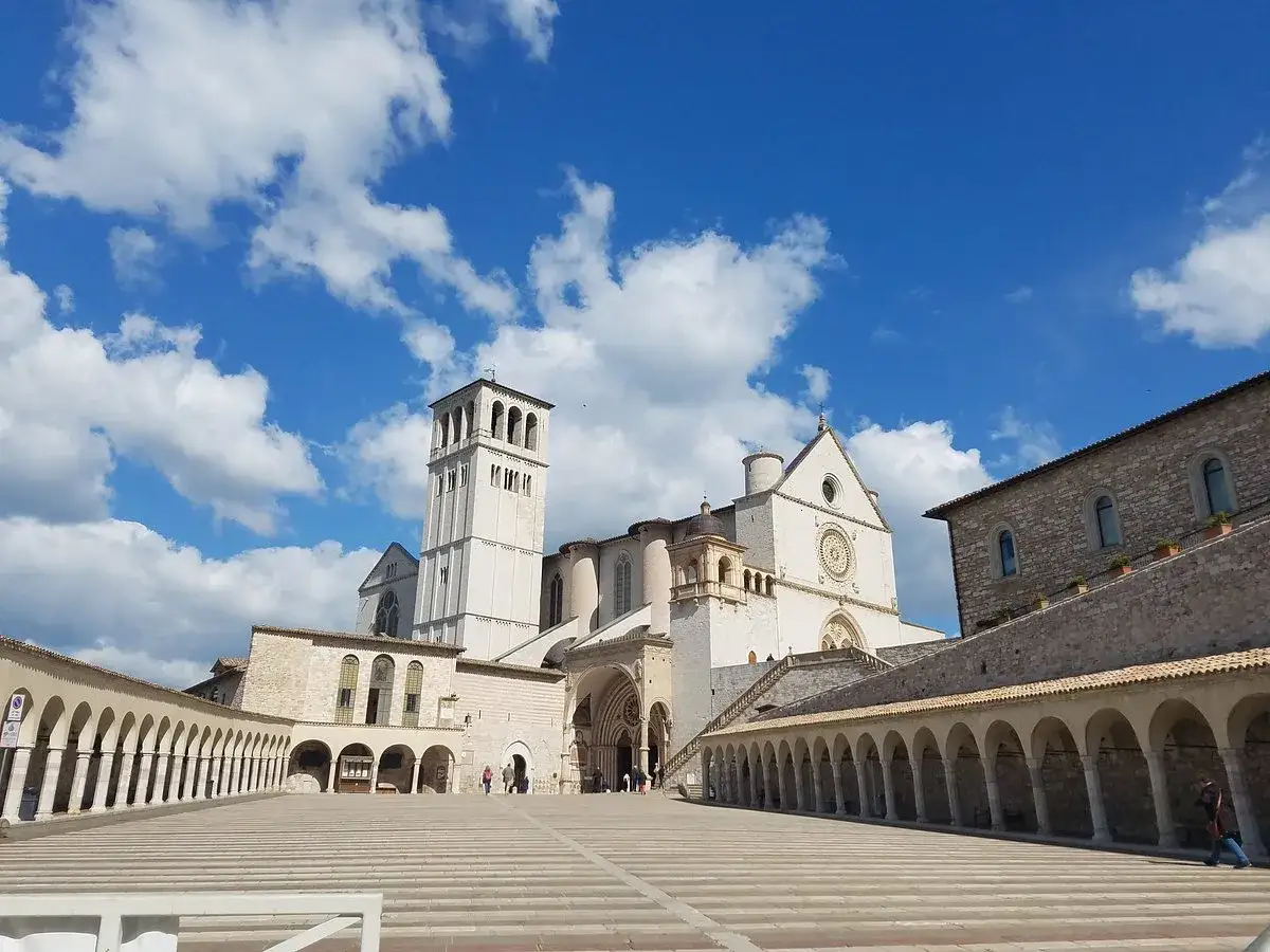 Piazza del centro storico di Assisi con la Basilica di San Francesco e il suo campanile sotto un cielo azzurro con nuvole bianche.