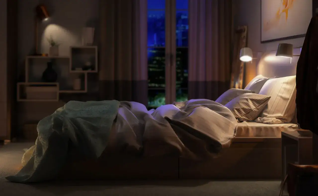 Un letto disfatto con una coperta azzurra, pronto per dormire fuori casa. La luce soffusa illumina la stanza, mentre fuori si intravede la citt&agrave; notturna.