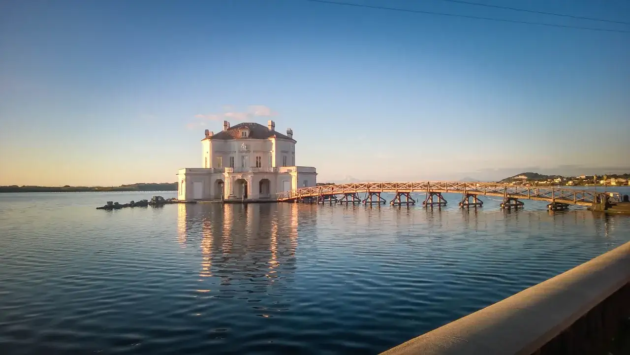 La casa di Pinocchio a Bacoli, un'elegante villa sull'acqua collegata da un ponte di legno, riflette la luce dorata del tramonto.