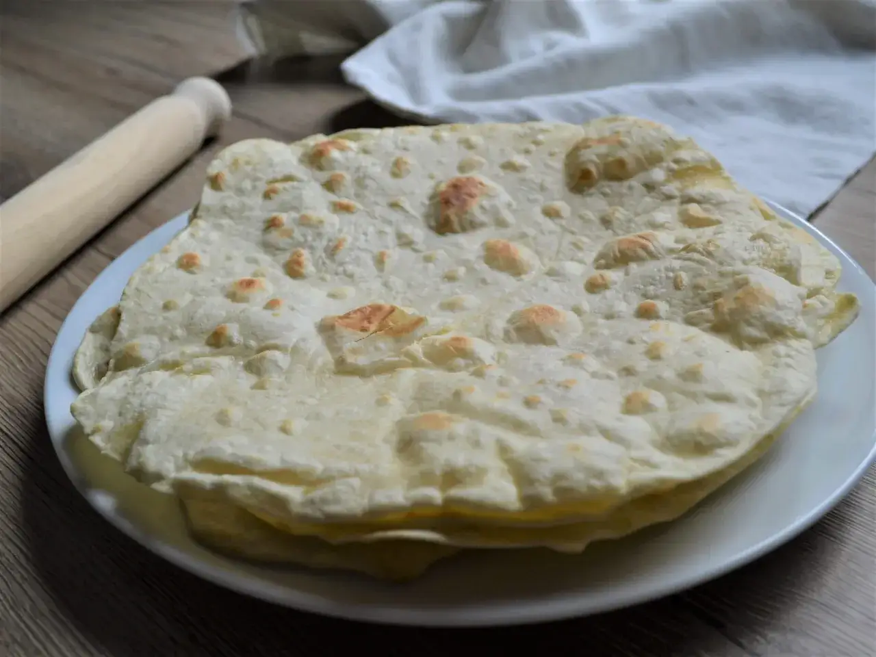 Impasto piadina appena cotto, soffice e leggermente dorato, pronto per essere farcito. Un mattarello e un canovaccio completano la scena.