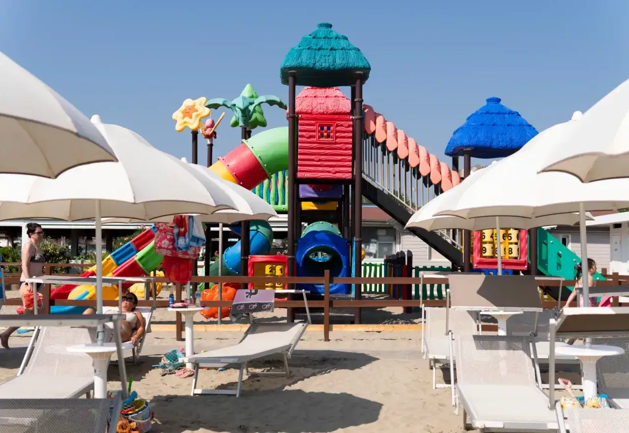 Area giochi colorata in un villaggio a Cesenatico, con scivoli, ombrelloni bianchi e lettini sulla sabbia.