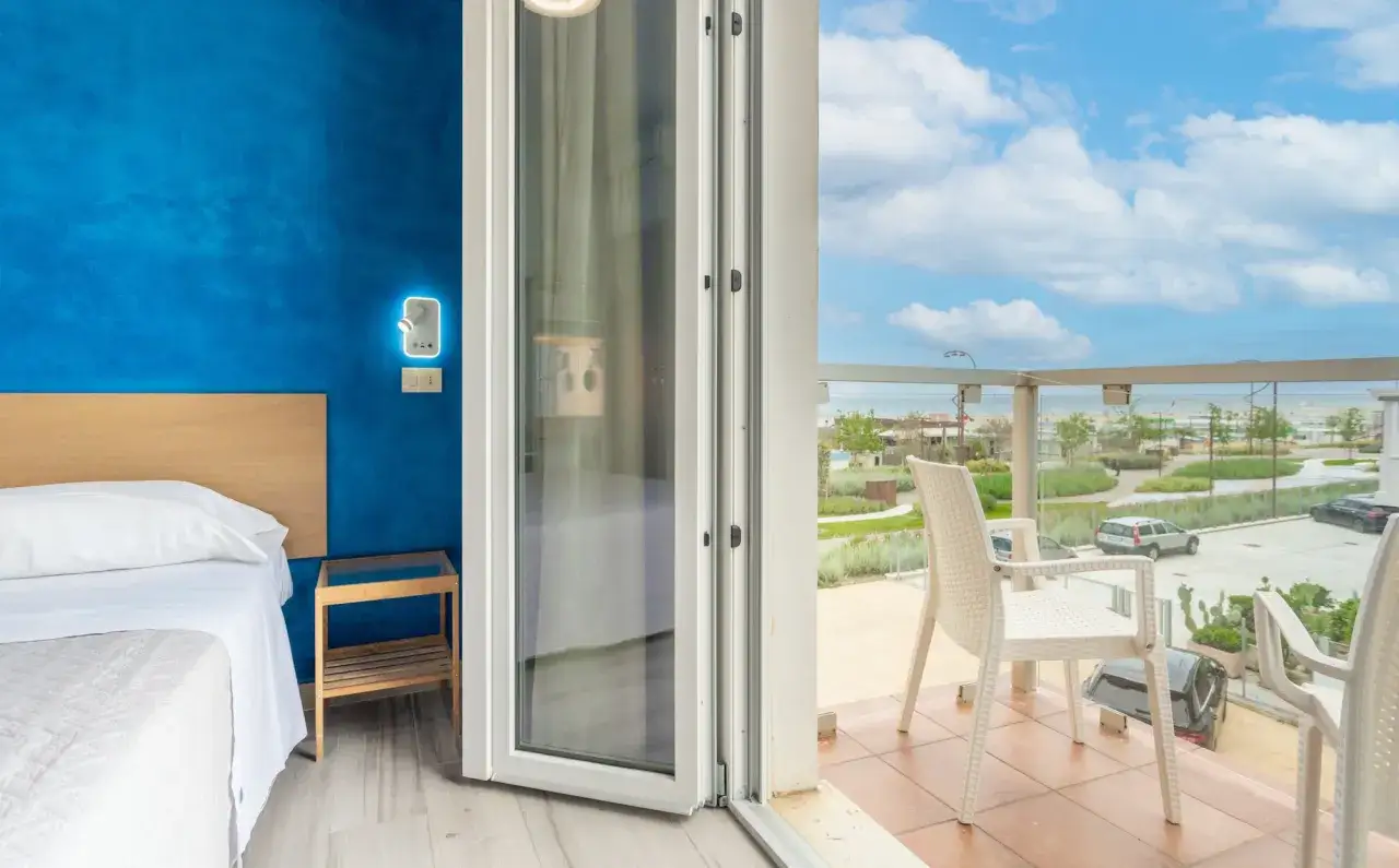 Camera con vista mare all'hotel Tamanco. Letto bianco, parete blu acceso e balcone con sedie bianche.