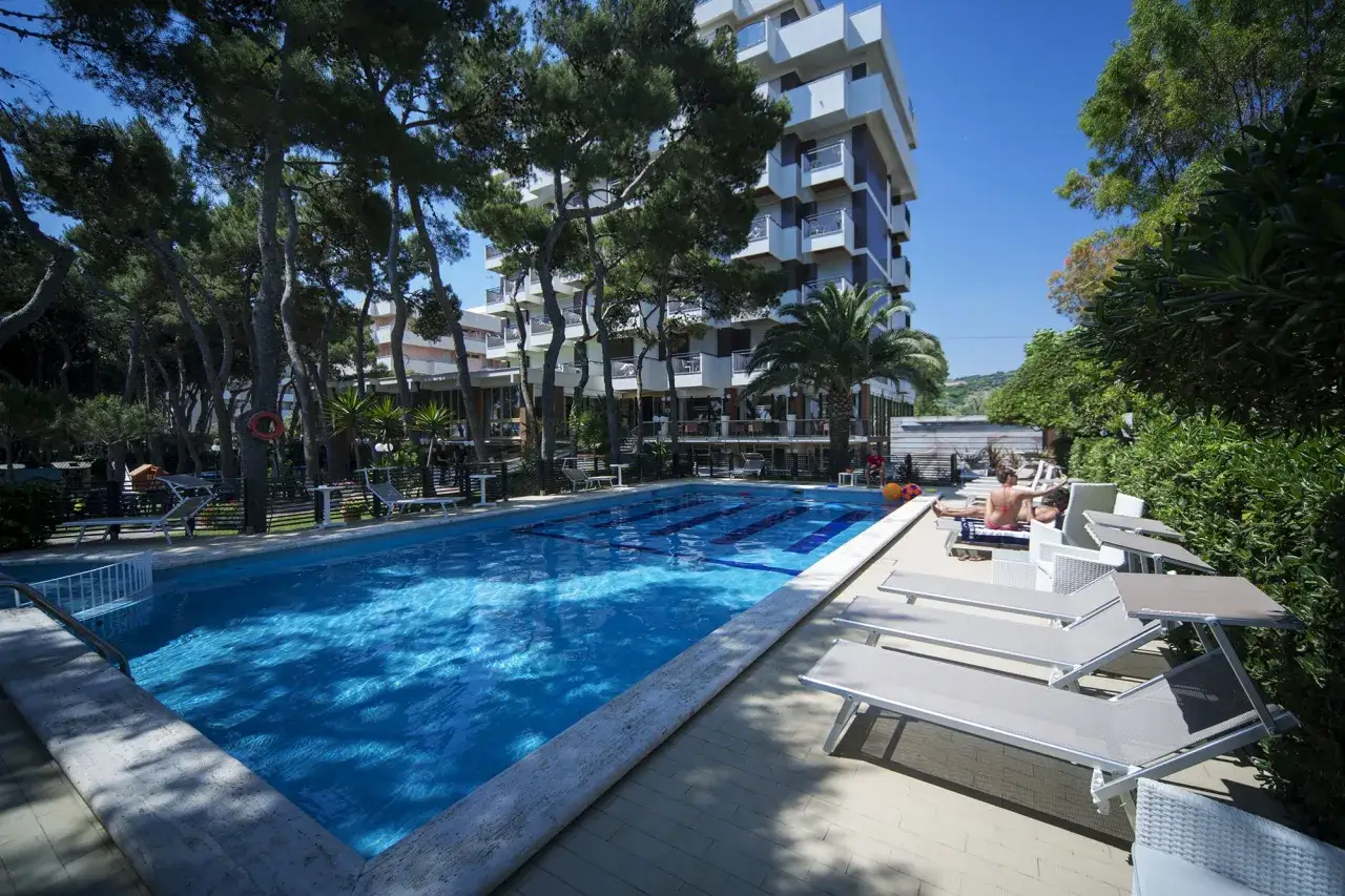 Piscina e lettini prendisole all'esterno del promenade hotel Giulianova, con alberi e edificio sullo sfondo.