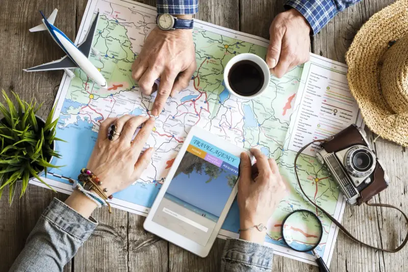 Pianificare viaggi senza agenzia: mani che studiano una mappa, un tablet con sito di viaggi, aereo giocattolo e macchina fotografica vintage.