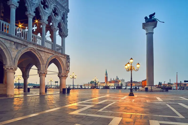 L'alba illumina il colonnato del Palazzo Ducale, proiettando lunghe ombre sulla piazza. In lontananza, il leone alato veglia su Piazza San Marco.